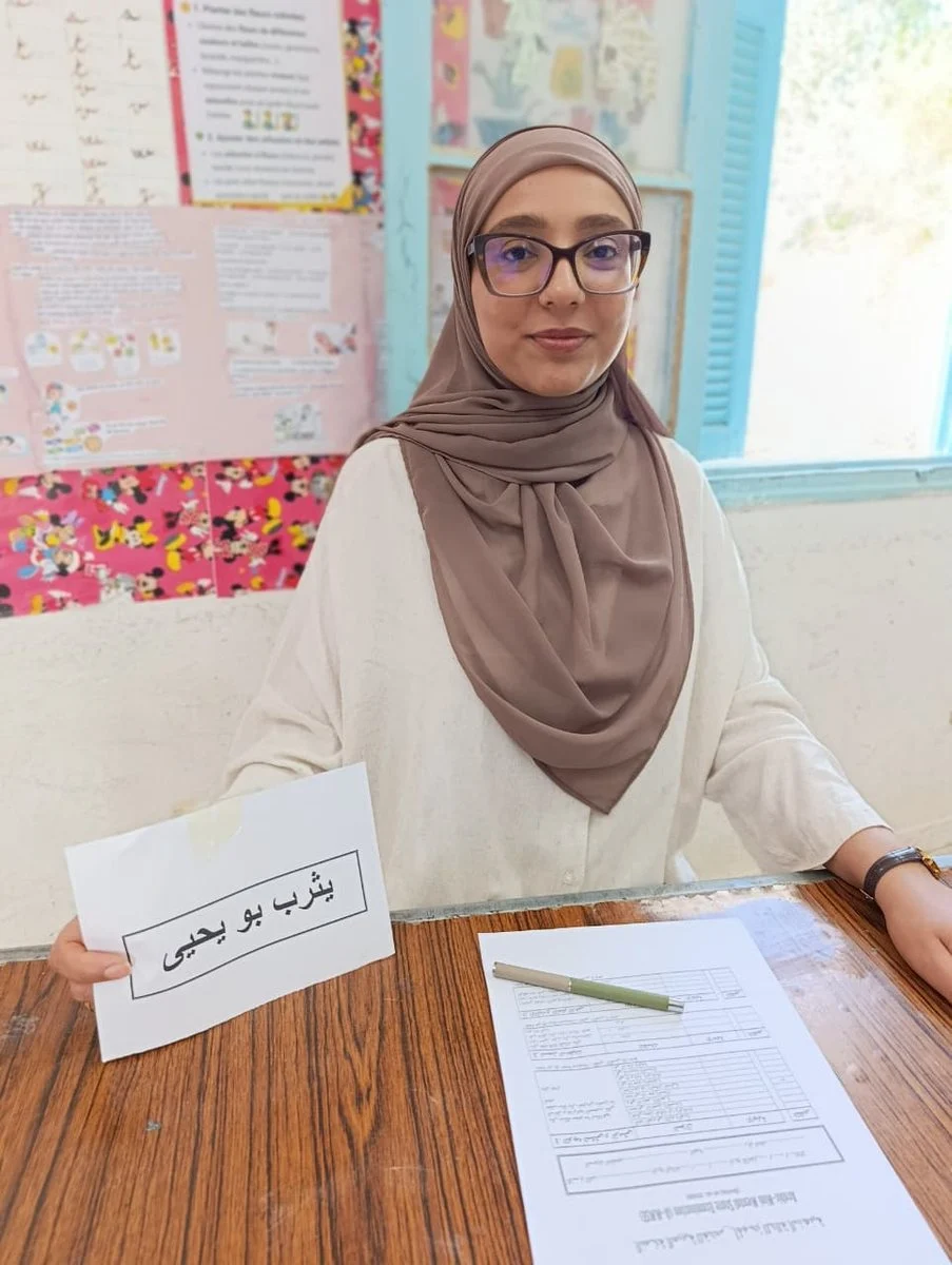 امرأة محجبة جالسة إلى طاولة استقبال داخل قاعة وأمامها أوراق ولافتة صغيرة