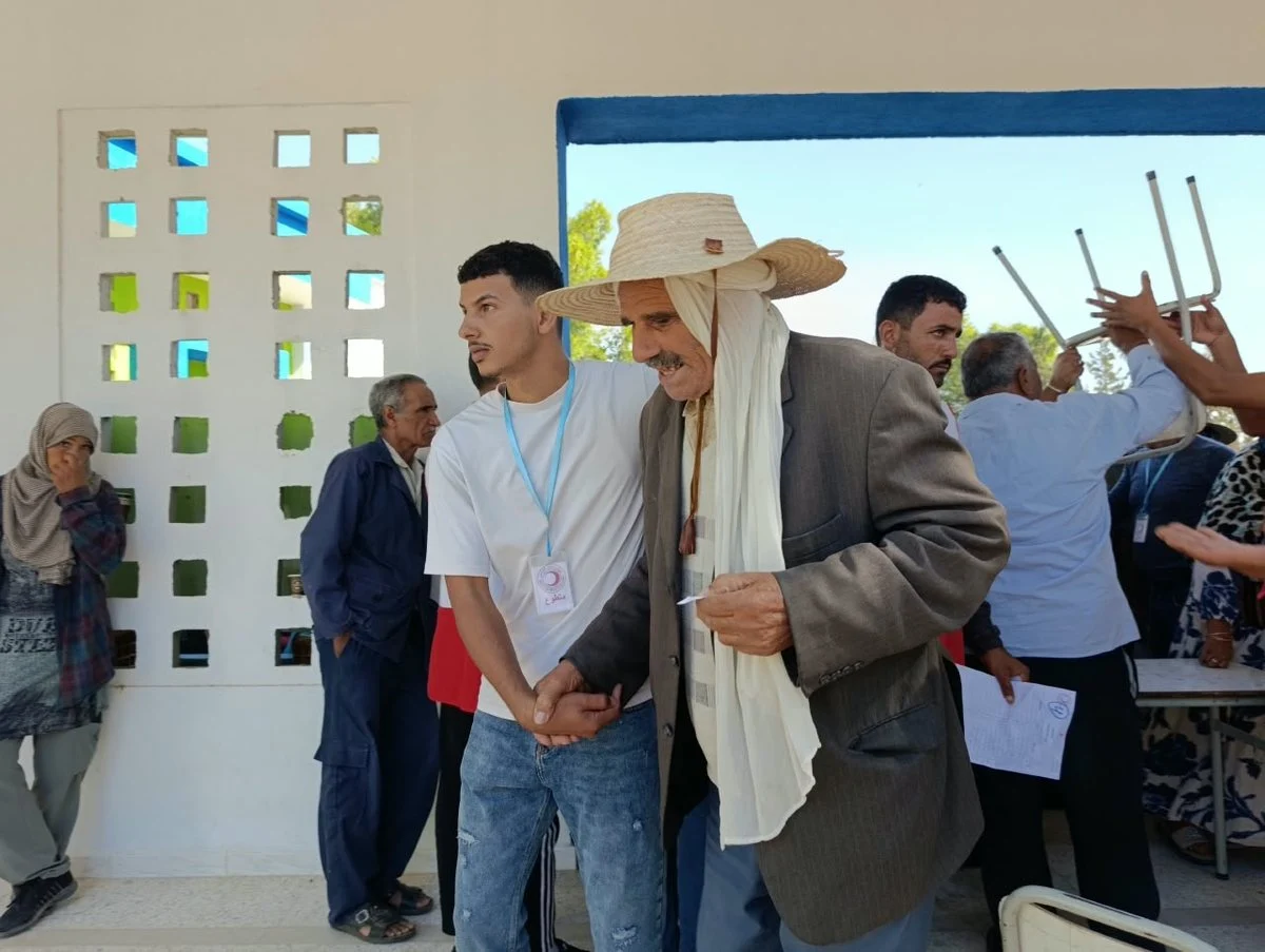 شاب يمسك بذراع رجل مسنّ يرتدي قبعة من القش وسط حضور داخل رواق بسيدي بوبكر