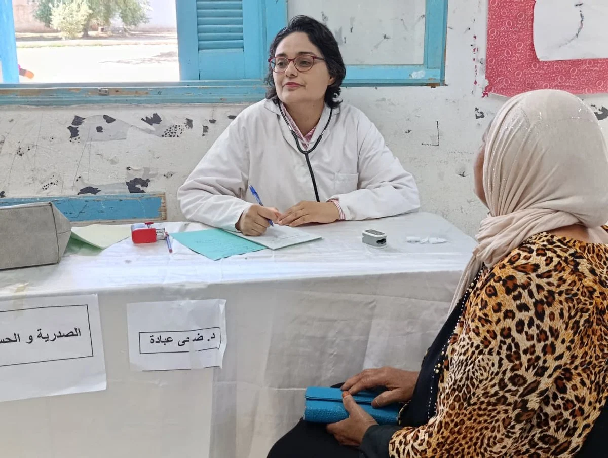طبيبة بمئزر أبيض وسماعة طبية تدوّن ملاحظات وأمامها امرأة مسنّة جالسة، وعلى الطاولة لافتة اختصاص طبي