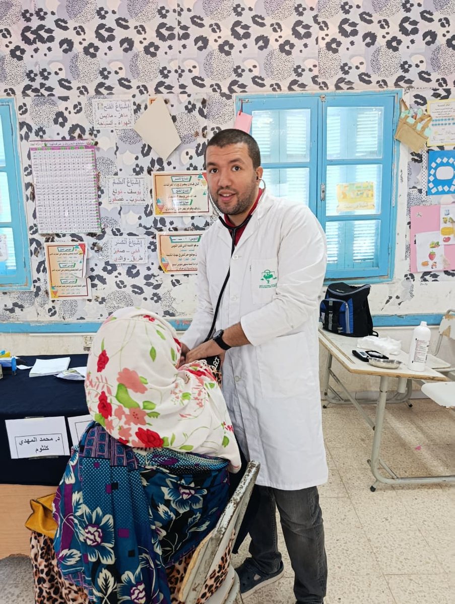 طبيب بمئزر أبيض يقيس ضغط الدم لامرأة مسنّة بحجاب مزهّر وبجانبه جهاز قياس الضغط