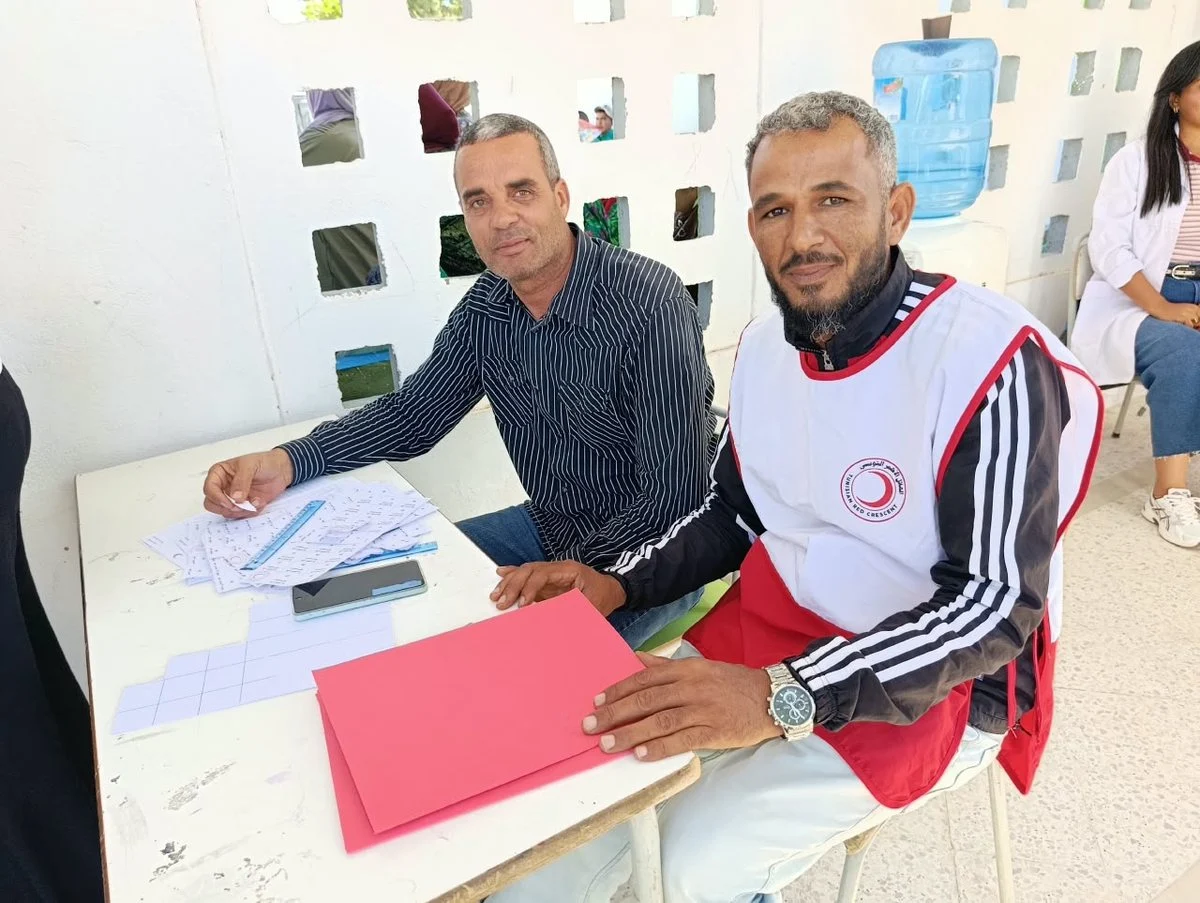 متطوع بسترة الهلال الأحمر جالس إلى طاولة بجانب رجل آخر وأمامهما أوراق وملف أحمر