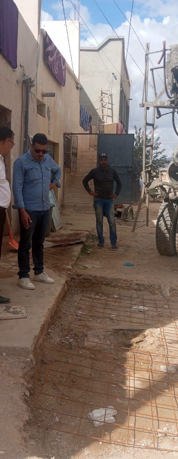 مسؤولان يعاينان حفرة مجهّزة بشبكة حديد في زقاق سكني بمنطقة العقيلة، وشاحنة خلّاطة على الجانب
