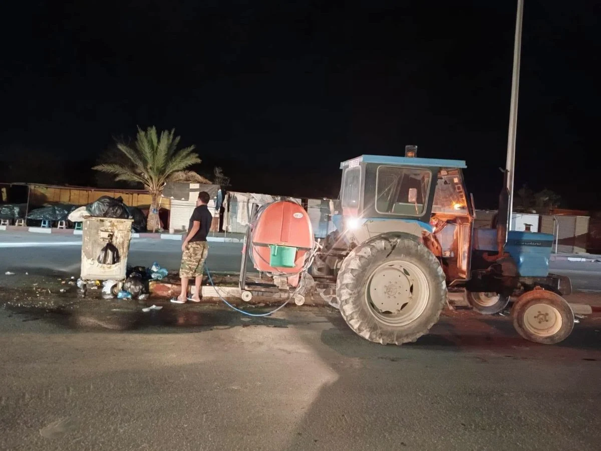 عامل بلدي يرش محيط حاوية فضلات قرب نخلة وأكشاك مغلقة بجانب جرار أزرق مجهز بخزان مداواة
