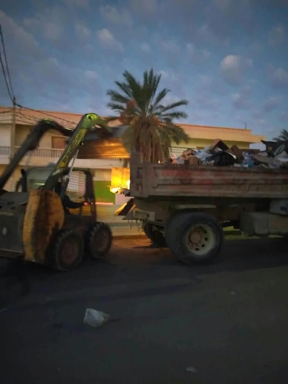 رافعة صغيرة تفرغ حمولتها في شاحنة قلابة مليئة بالفضلات عند الفجر في قفصة