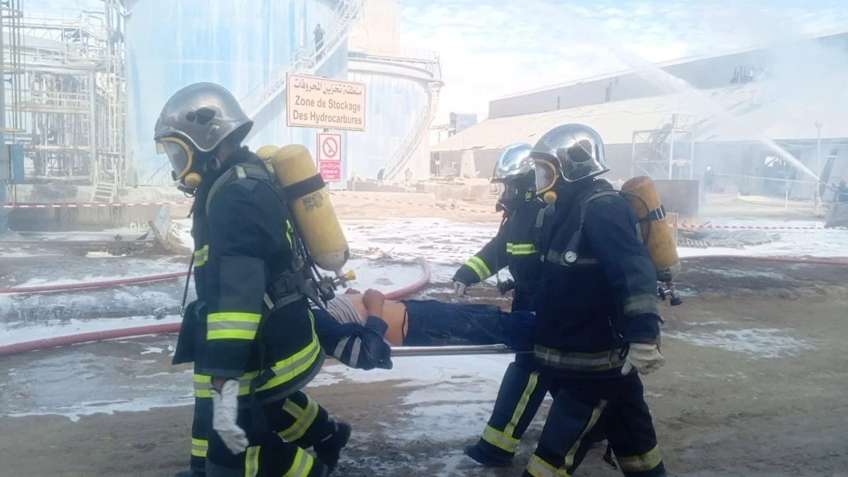 ثلاثة أعوان إطفاء بأجهزة تنفّس ينقلون مصابا على نقّالة أمام لافتة منطقة تخزين المحروقات بالمظيلة