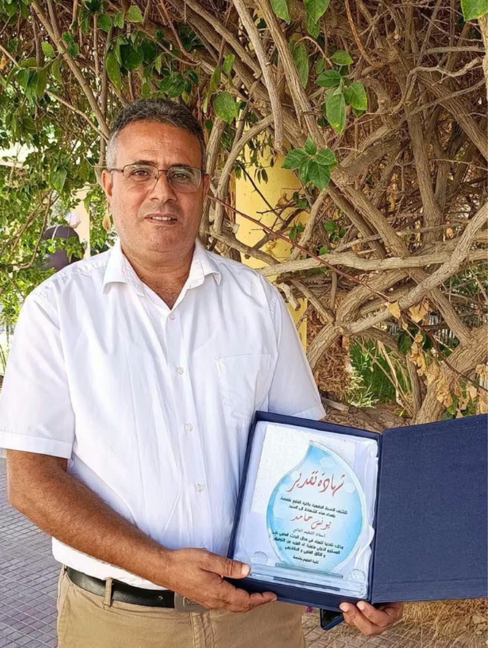 الأستاذ يونس حامد يحمل شهادة تقدير زجاجية على شكل قطرة ماء من كلية العلوم بقفصة تحت أغصان شجرة متشابكة