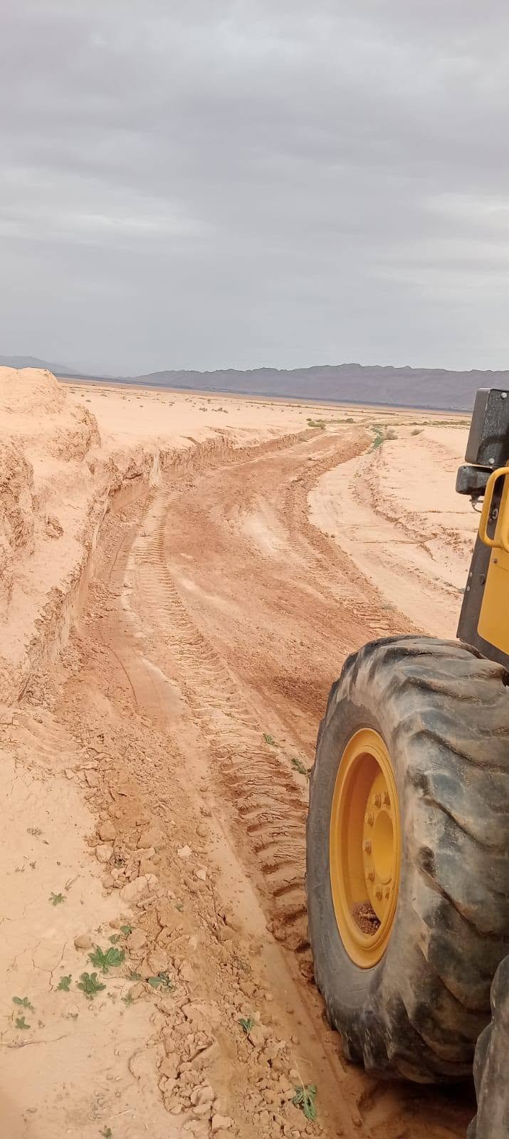 مسلك ترابي متعرّج بين حواف رملية تحت سماء غائمة وعجلة آلة تسوية في المقدمة