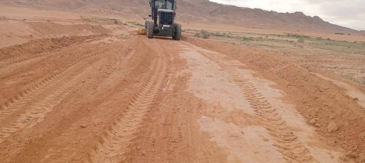 آلة تسوية تعمل على مسلك ترابي أحمر في سهل تحيط به الجبال