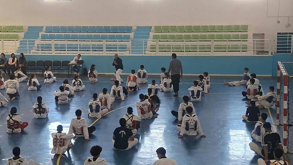 مدرّب يشرح حركة أمام لاعبين جالسين على الأرض بصدرياتهم الواقية، والمدارج الخضراء والزرقاء في الخلفية