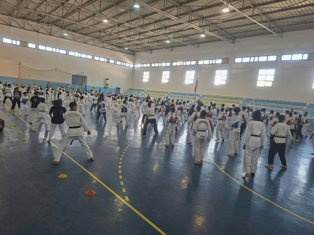 لاعبو تايكوندو بأحزمة ملوّنة يؤدّون حركات جماعية على أرضية القاعة الزرقاء تحت السقف المعدني