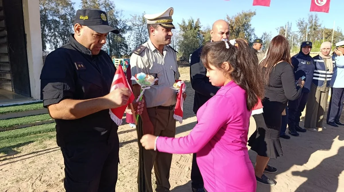فتاة بقميص وردي تقدّم علما صغيرا ووردة لعونين من الأمن بزيّ رسمي في ساحة جمعية التفاؤل بقفصة