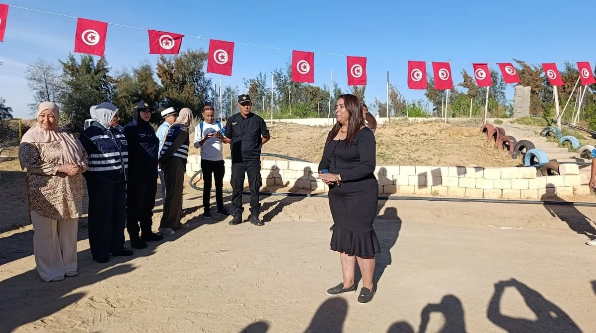 امرأة بفستان أسود تتحدث في ساحة جمعية التفاؤل بقفصة أمام متطوعات بسترات زرقاء وعون أمن تحت أعلام تونسية
