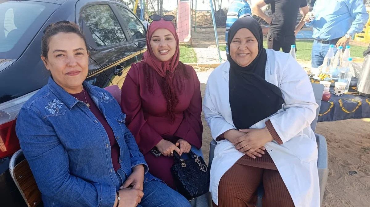 ثلاث نساء جالسات مبتسمات، إحداهن بمئزر أبيض، خلال الاحتفال في الهواء الطلق بجمعية التفاؤل بقفصة