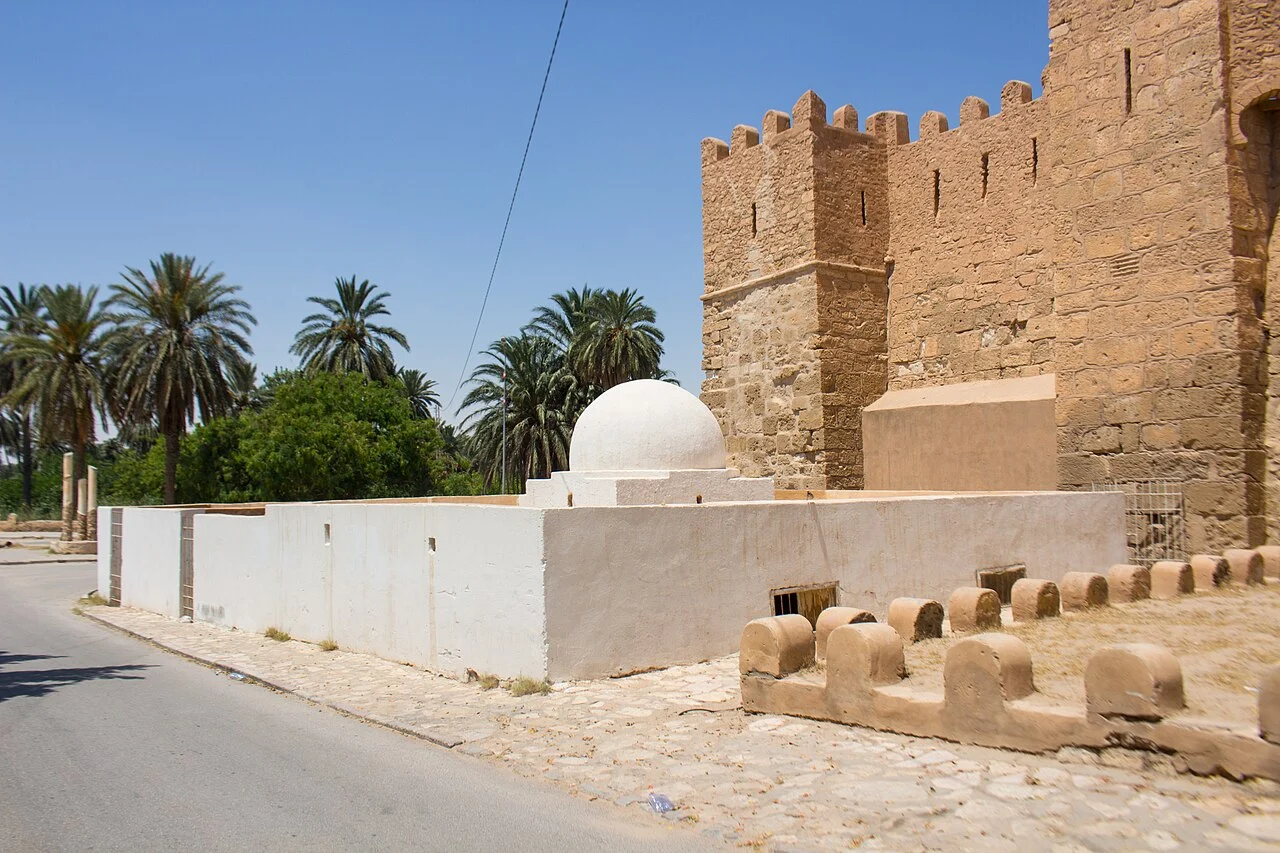 مبنى الترميل بقفصة من الخارج بقبّته البيضاء وجدار القصبة الحجري خلفه ونخيل الواحة