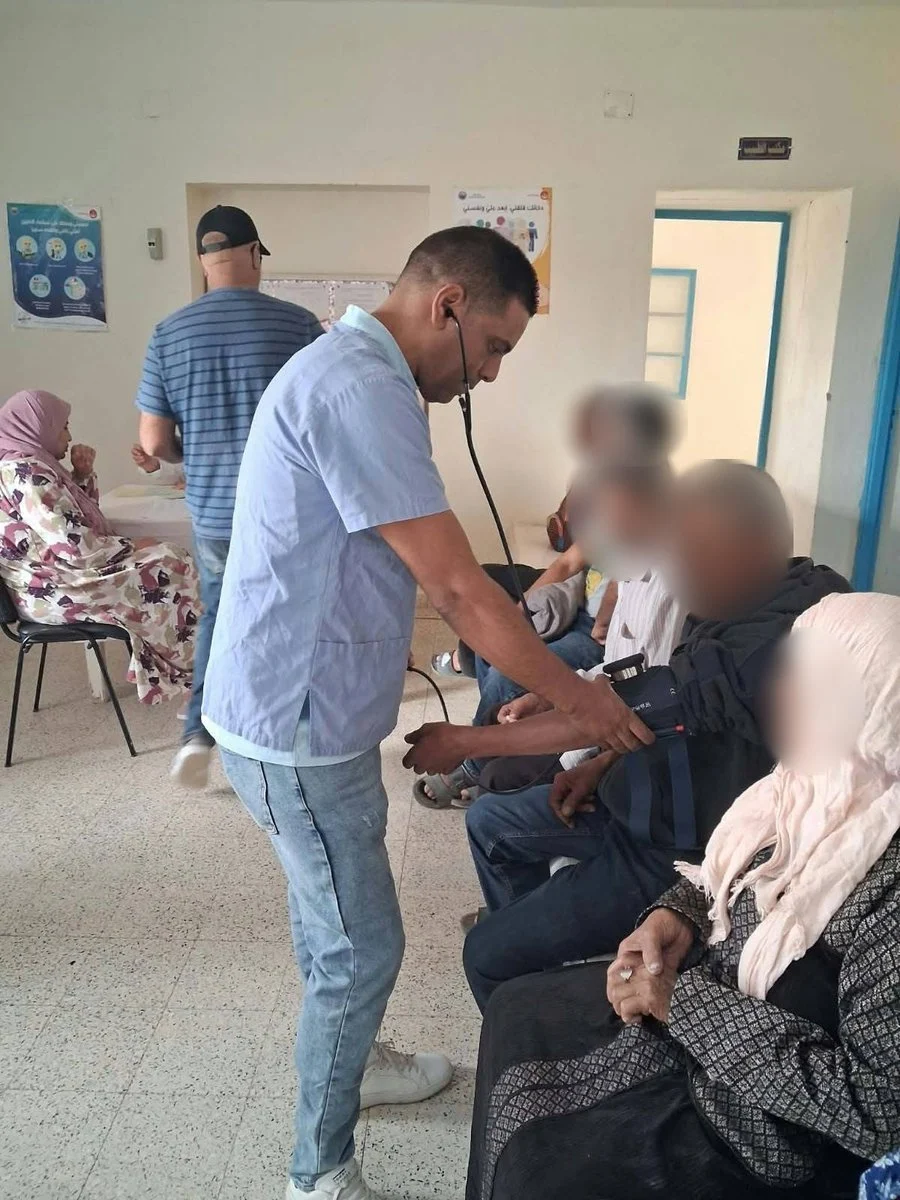 ممرض يقيس ضغط الدم لرجل مسنّ في قاعة انتظار المستوصف بينما ينتظر آخرون دورهم