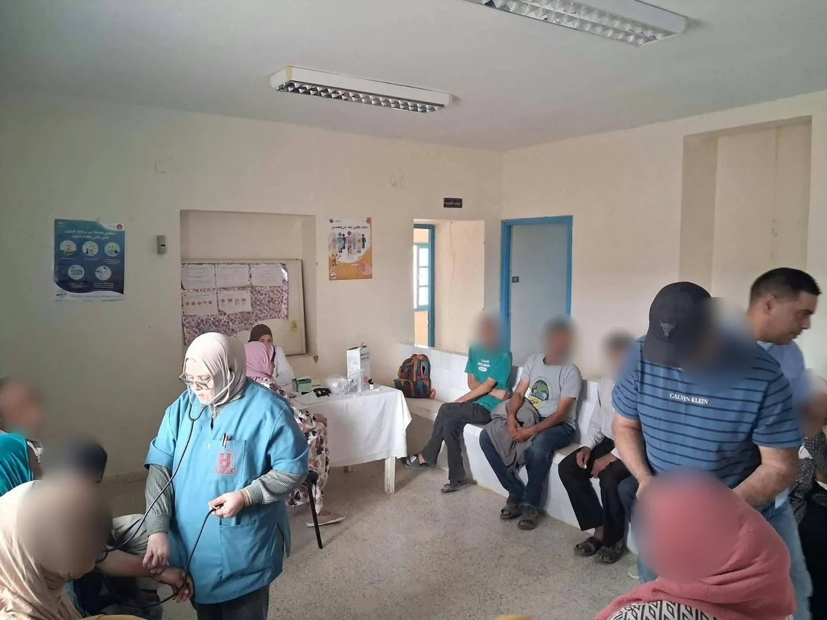 ممرضة تفحص امرأة بالسماعة الطبية ورجال ينتظرون على مقعد قاعة المستوصف