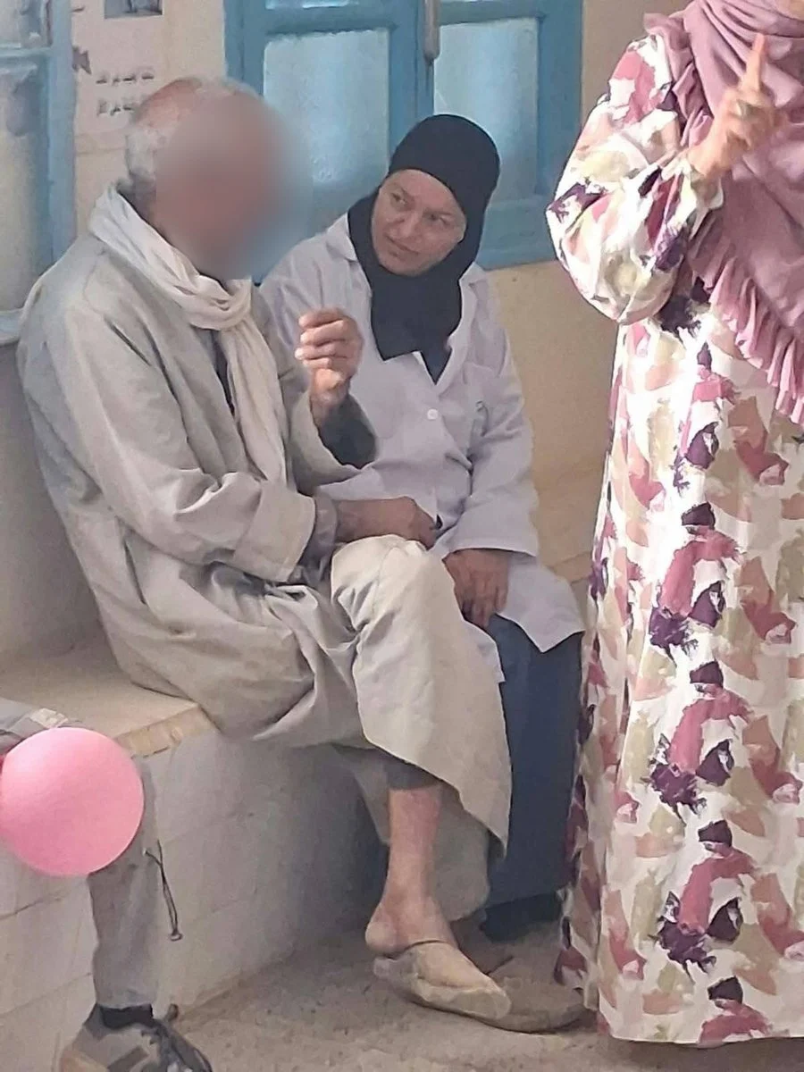 ممرضة تصغي إلى رجل مسنّ يتحدّث وهو جالس على مقعد حجري في المستوصف
