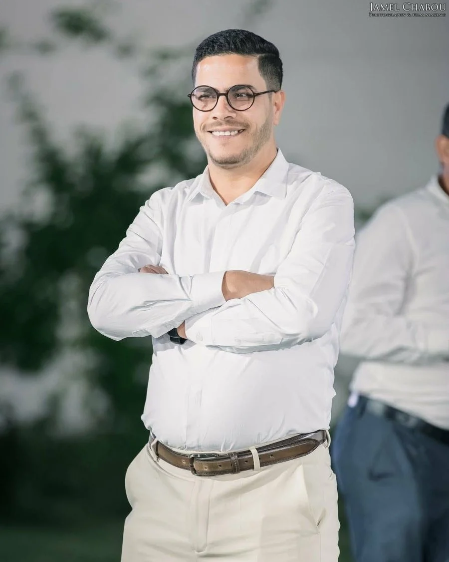 ماهر بن ظاهر، المكلف بالإعلام، مبتسما بنظارات دائرية وقميص أبيض وذراعاه معقودتان
