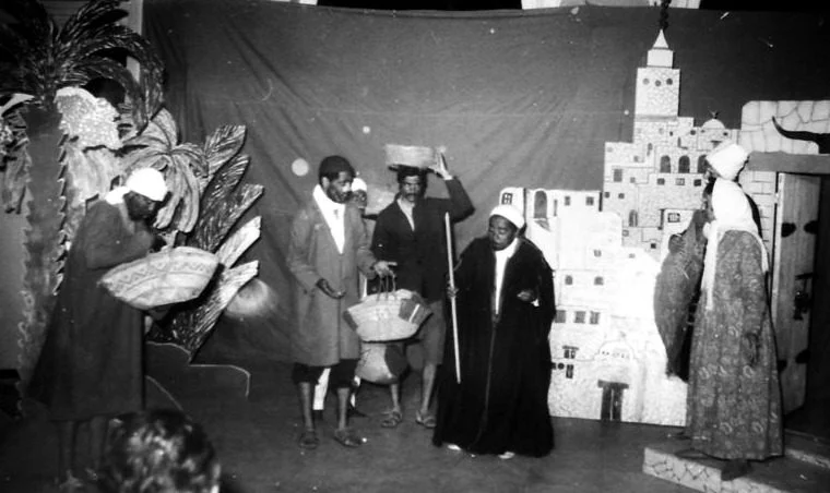 Scène en noir et blanc d'un spectacle, avec un décor de ville surmonté d'un minaret et des comédiens en costumes traditionnels