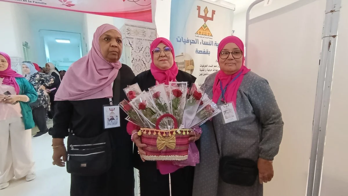 ثلاث مشاركات يحملن سلة ورود حمراء أمام لافتة جمعية النساء الحرفيات بقفصة