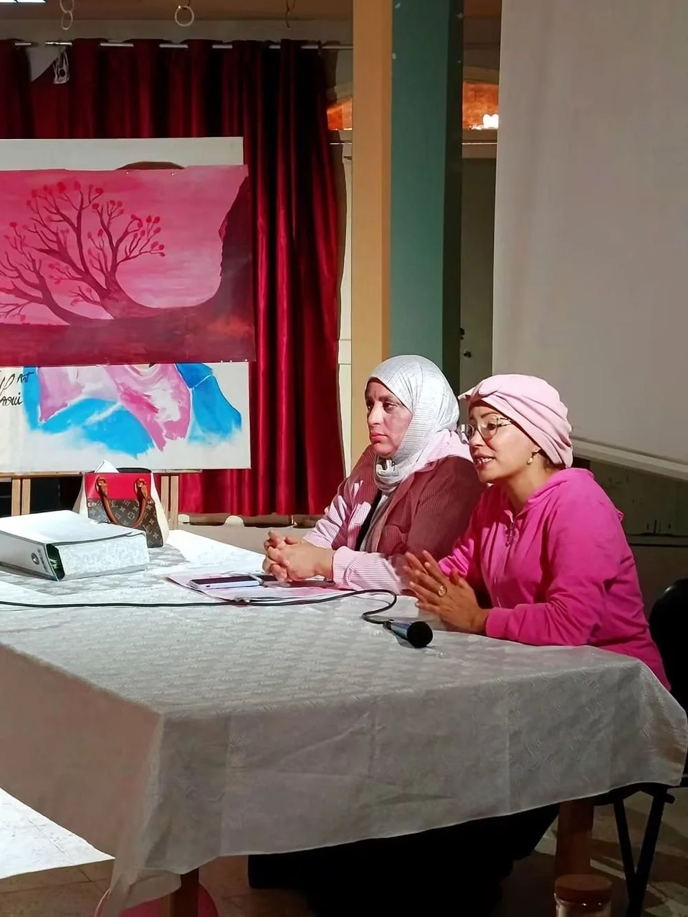 متدخلتان إلى طاولة خلال جلسة توعوية، وخلفهما لوحة مرسومة لشجرة بالألوان الوردية وستار أحمر