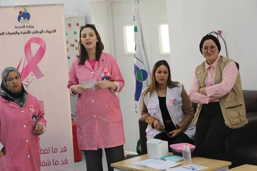 قابلات بمآزر وردية يقدّمن إرشادات صحية أمام لافتة الديوان الوطني للأسرة والعمران البشري حول سرطان الثدي