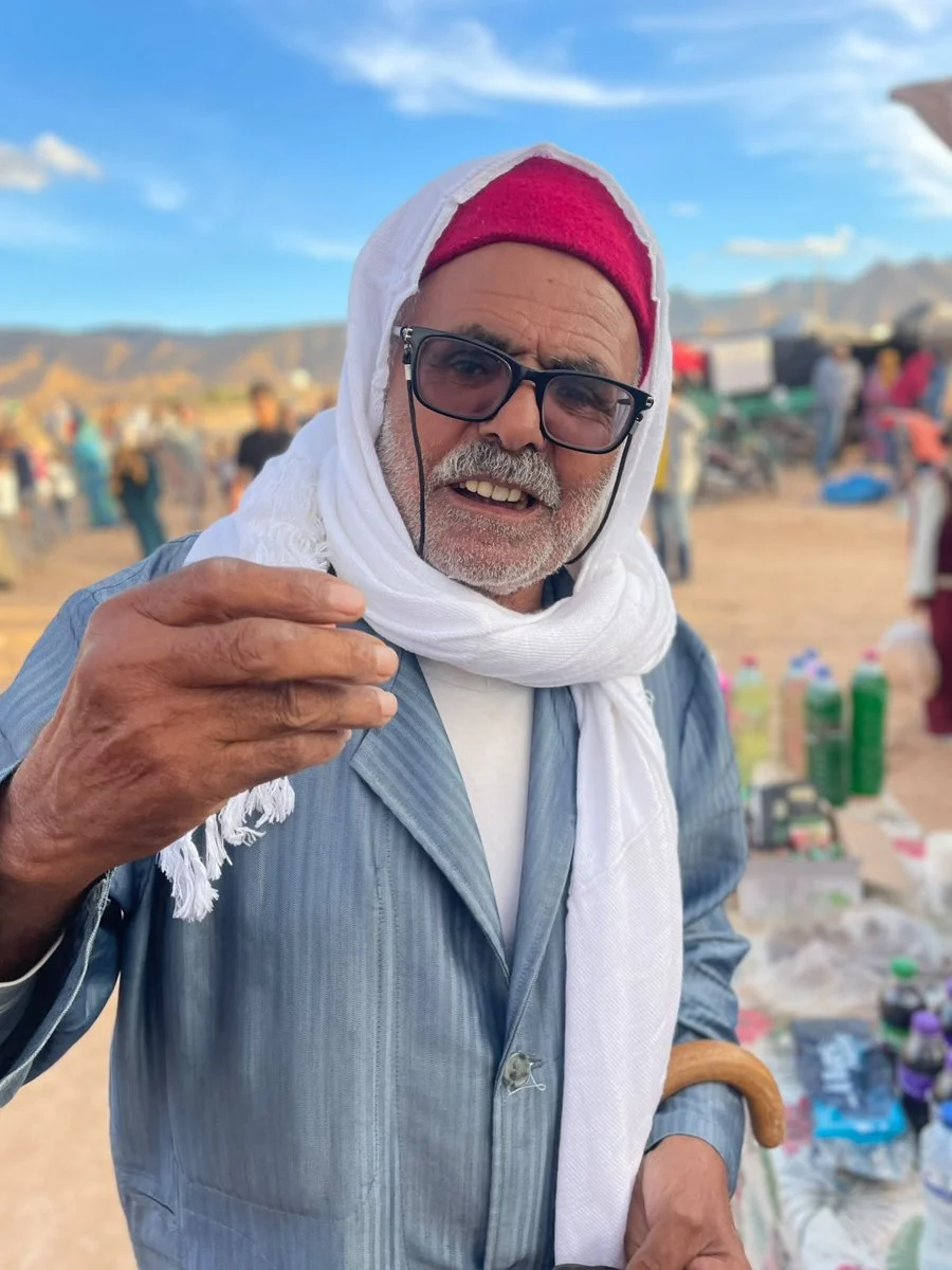 رجل مسنّ بشاشية حمراء ولحاف أبيض يتحدّث في فضاء مهرجان صوت الجبل وخلفه جبال أم العلق