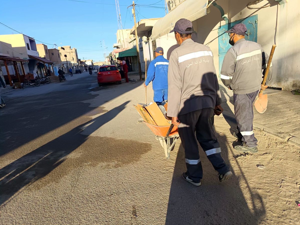 أعوان بلدية أم العرائس يدفعون عربة يدوية محمّلة بالكراتين على جانب أحد الشوارع