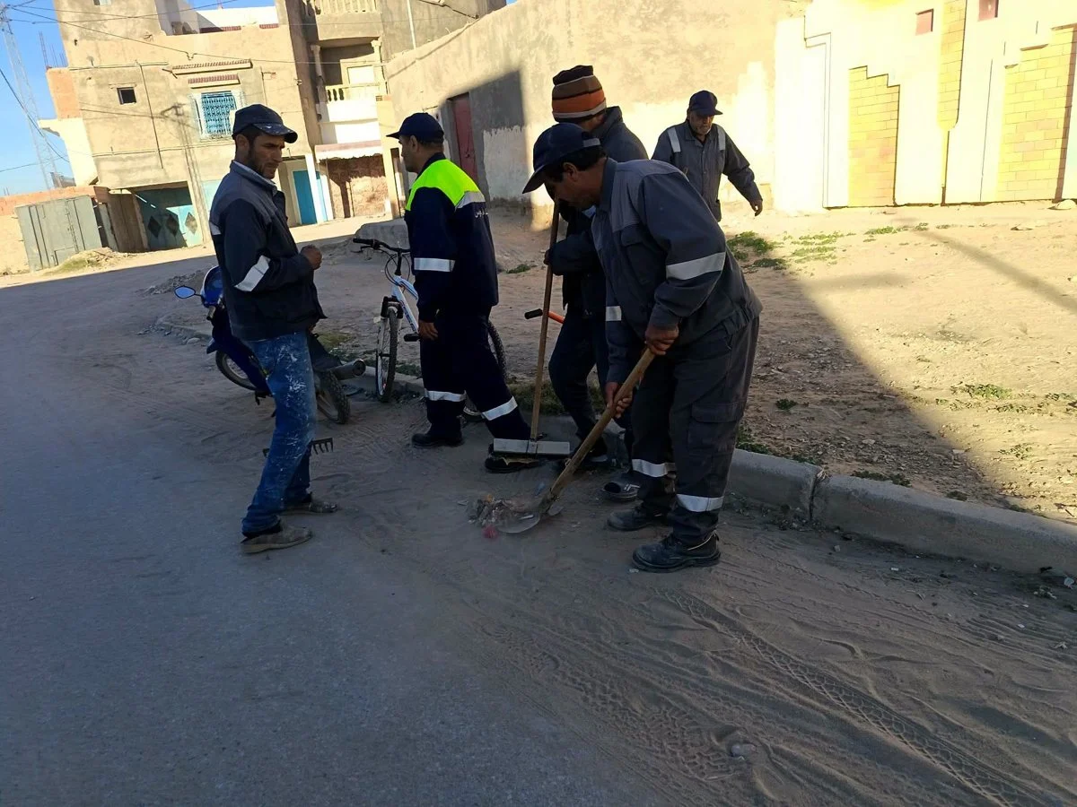 مجموعة من أعوان النظافة يكنسون الأتربة والفضلات على حافة رصيف بأحد أحياء أم العرائس