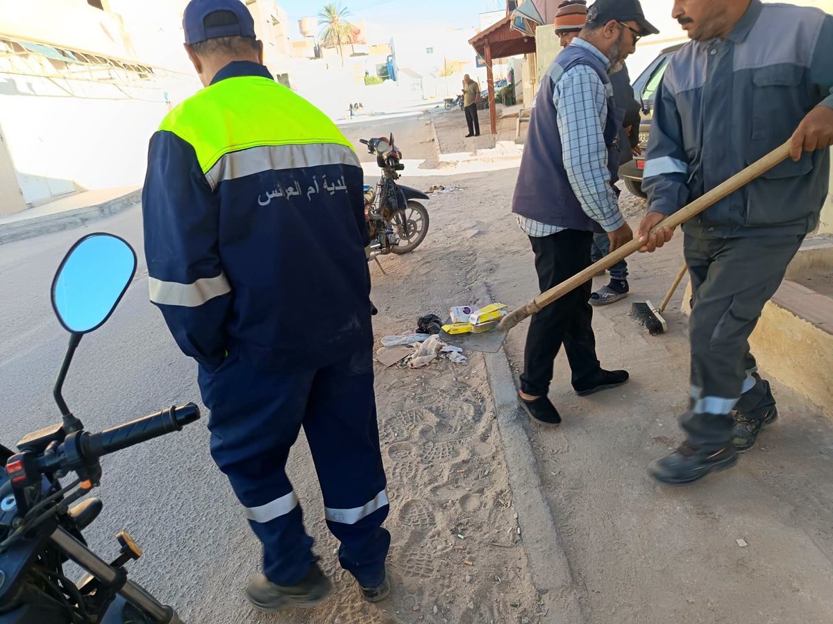 عون نظافة يجرف فضلات متراكمة على حافة رصيف قرب دراجة نارية بأم العرائس