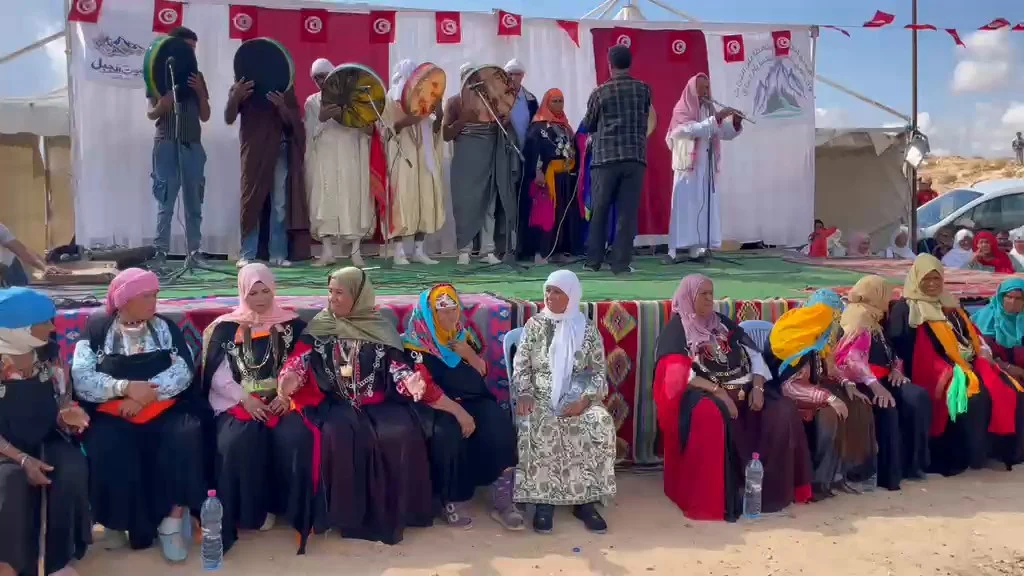 نساء مسنّات بلباس تقليدي وحليّ يجلسن أمام منصّة مهرجان صوت الجبل وعليها عازفو البندير والقصبة