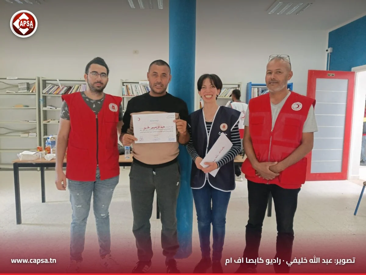 مشارك بقميص أسود يرفع شهادة مشاركته بين متطوعَين من الهلال الأحمر ومنسّقة الدورة