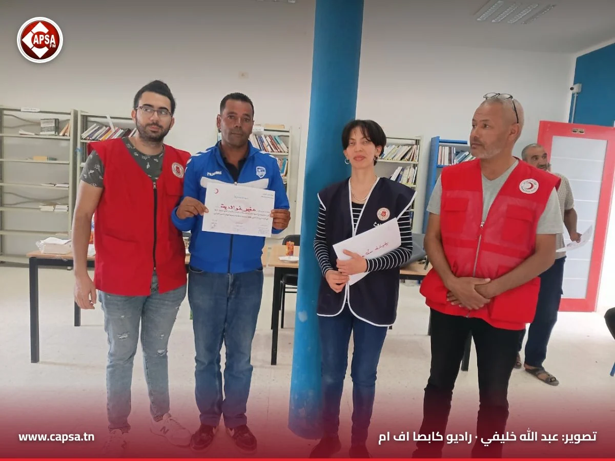 مشارك بسترة رياضية زرقاء يعرض شهادة المشاركة إلى جانب ثلاثة من مؤطّري الدورة