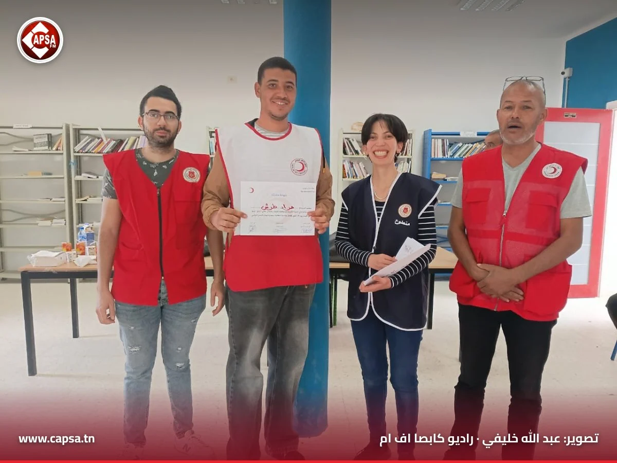 متدرّب مبتسم بصدرية الهلال الأحمر البيضاء يحمل شهادته بين المتطوعين في قاعة الدورة