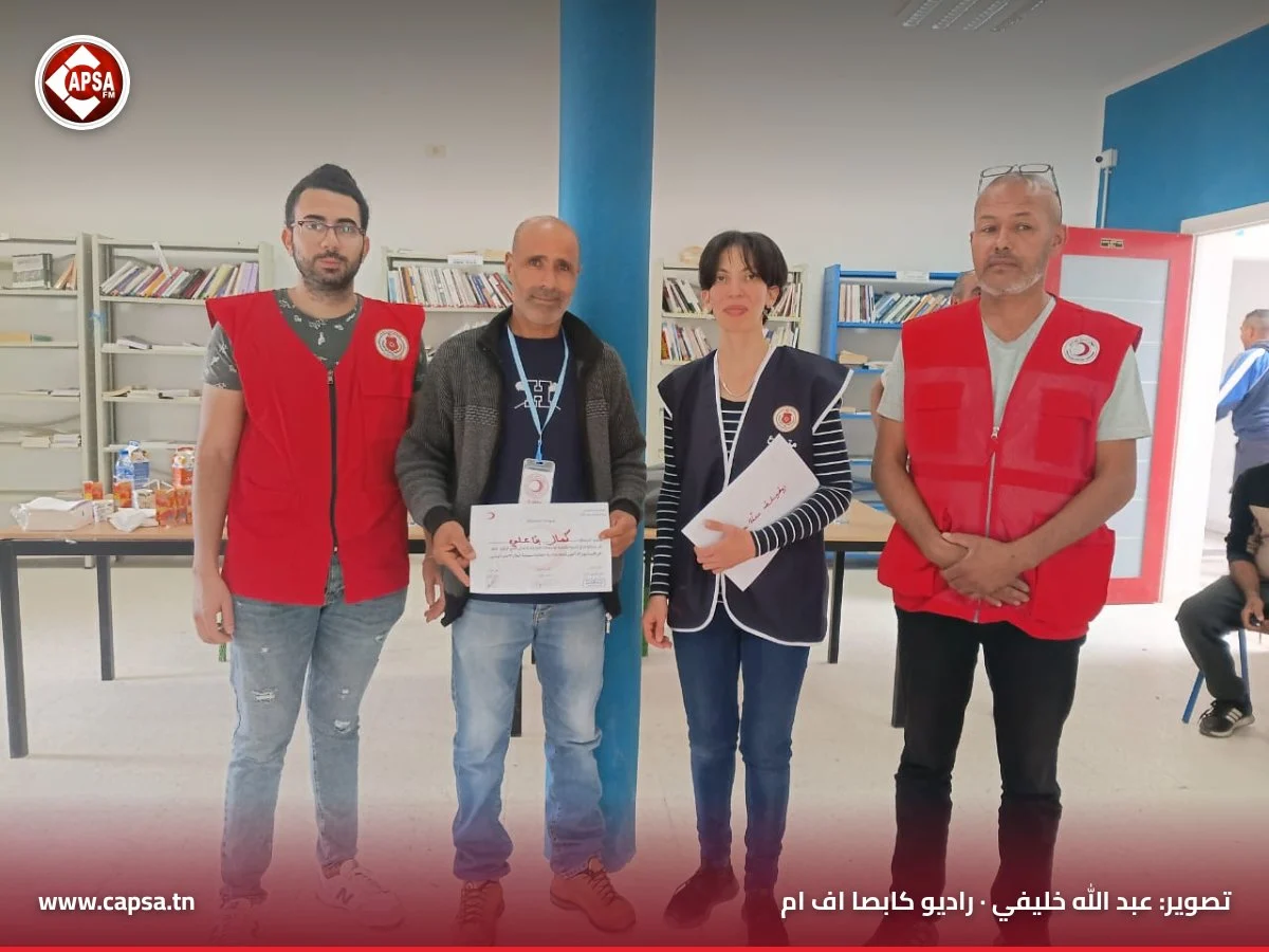 مشارك يحمل شارة الدورة وشهادة مشاركته واقفا بين متطوعَين ومنسّقة الدورة