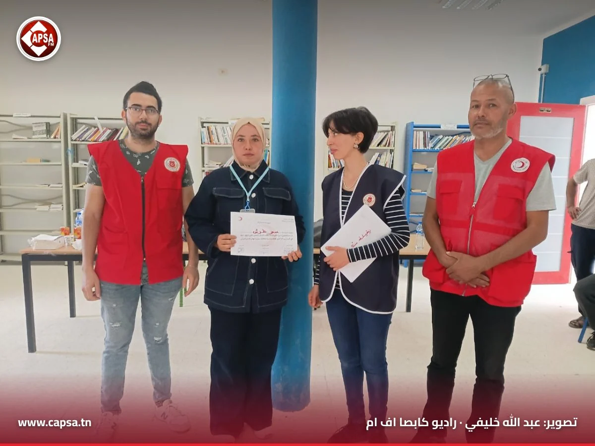 مشاركة بلباس أزرق داكن تعرض شهادتها في الإسعافات الأولية إلى جانب متطوعَين ومنسّقة الدورة