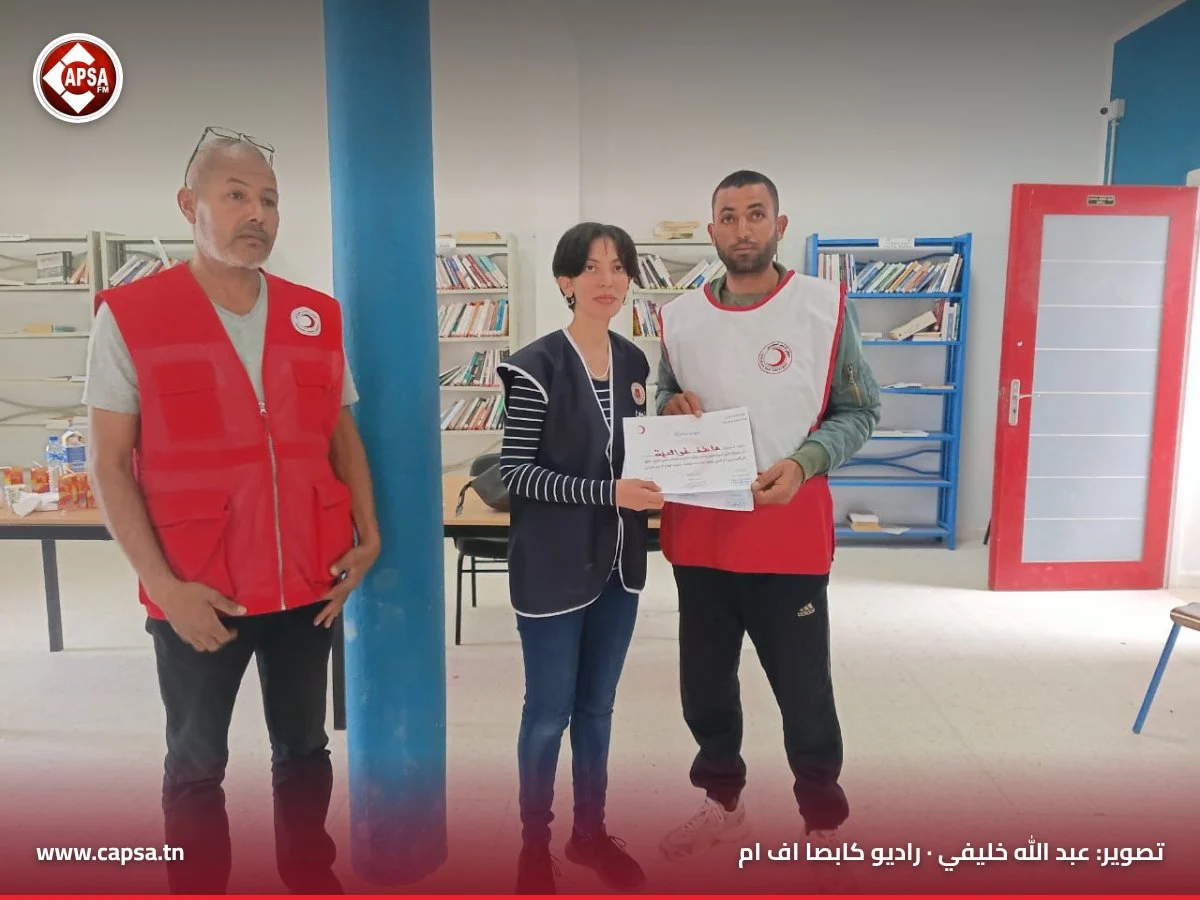 متطوعة تسلّم شهادة المشاركة لشاب يرتدي صدرية الهلال الأحمر البيضاء بحضور عضو من الفرع
