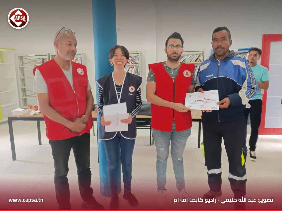متطوع من الهلال الأحمر يسلّم شهادة لمشارك يرتدي سترة رياضية زرقاء بحضور متطوعة وعضو من الفرع