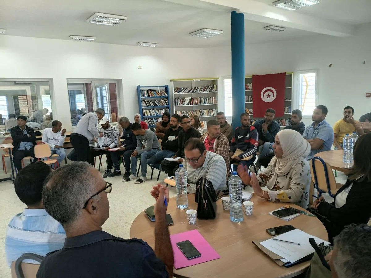 مشاركون في اليوم التحسيسي لدفع الاستثمار الخاص يجلسون حول طاولات في المكتبة العمومية بسيدي بوبكر أمام رفوف الكتب والعلم التونسي