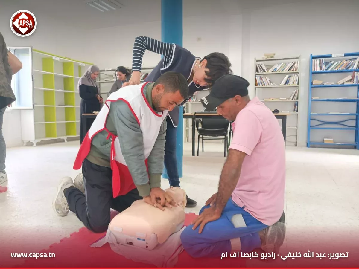 متدرّب يرتدي صدرية الهلال الأحمر يطبّق تدليك القلب على دمية تدريب بإشراف متطوعة، وإلى جانبه مشارك آخر في دورة سيدي بوبكر